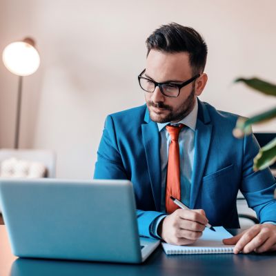 Handsome businessman in eyeglasses is making notes while working on a laptop.