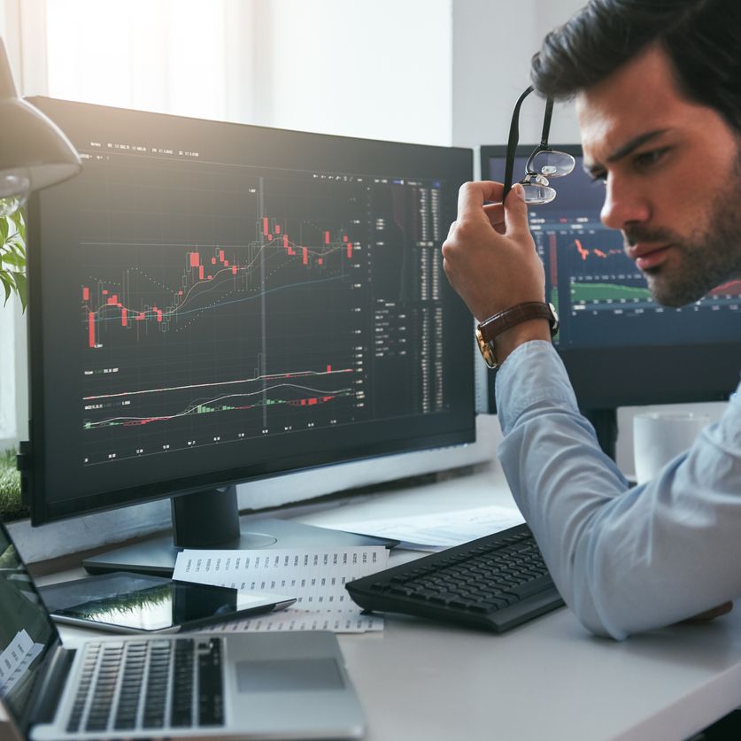 Successful investor. Serious young male trader holding eyeglasses and analyzing data and graphs on his laptop while working in his modern office. Stock exchange. Trade concept. Investment concept